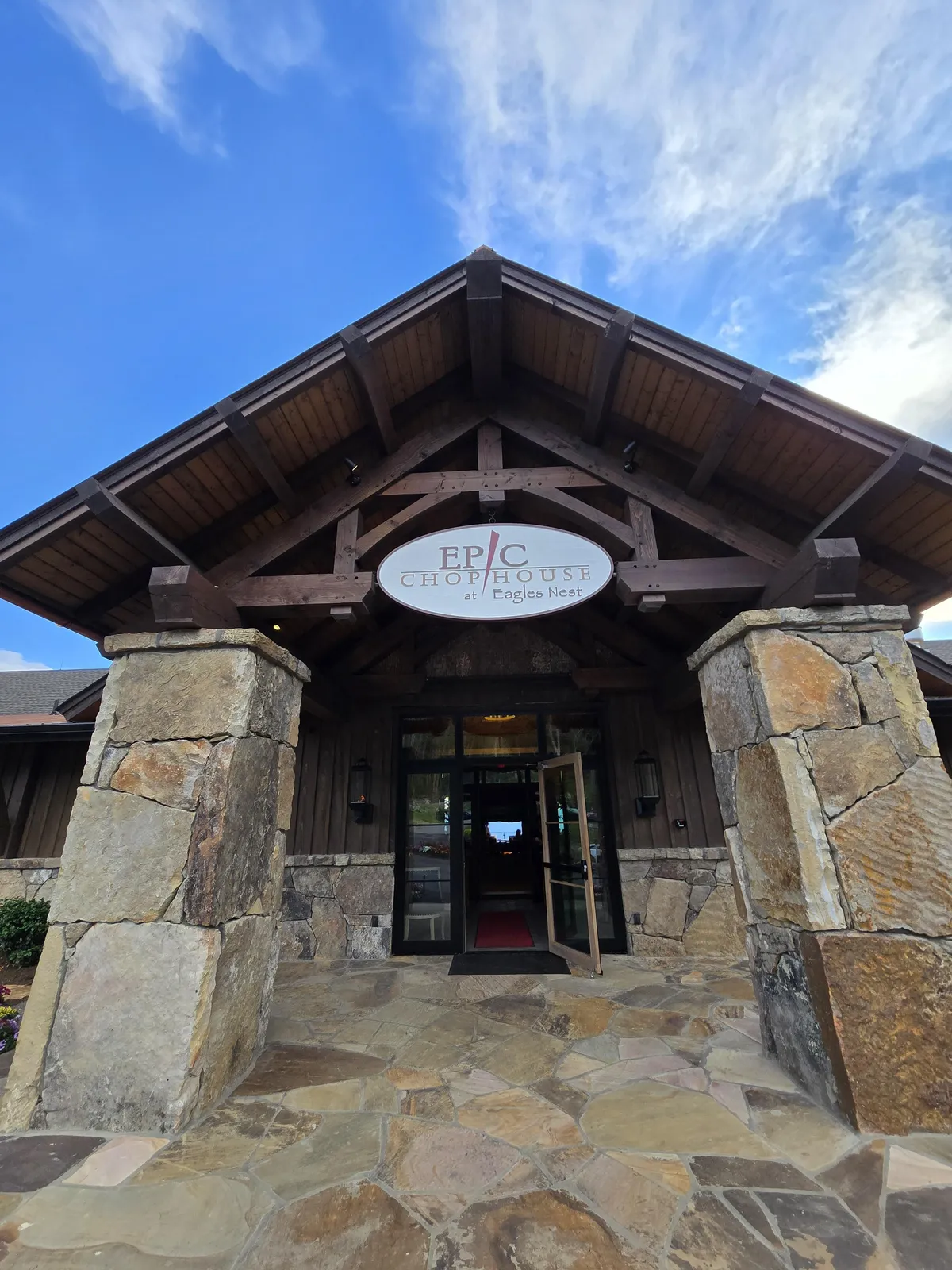 An inviting exterior view of the entrance to Epic Chophouse, featuring double doors, wooden beams, and stone columns with clear signage. The upscale architectural details and warm lighting make it feel exclusive and welcoming, aligning with the mention of resort amenities and fine dining within Eagles Nest.