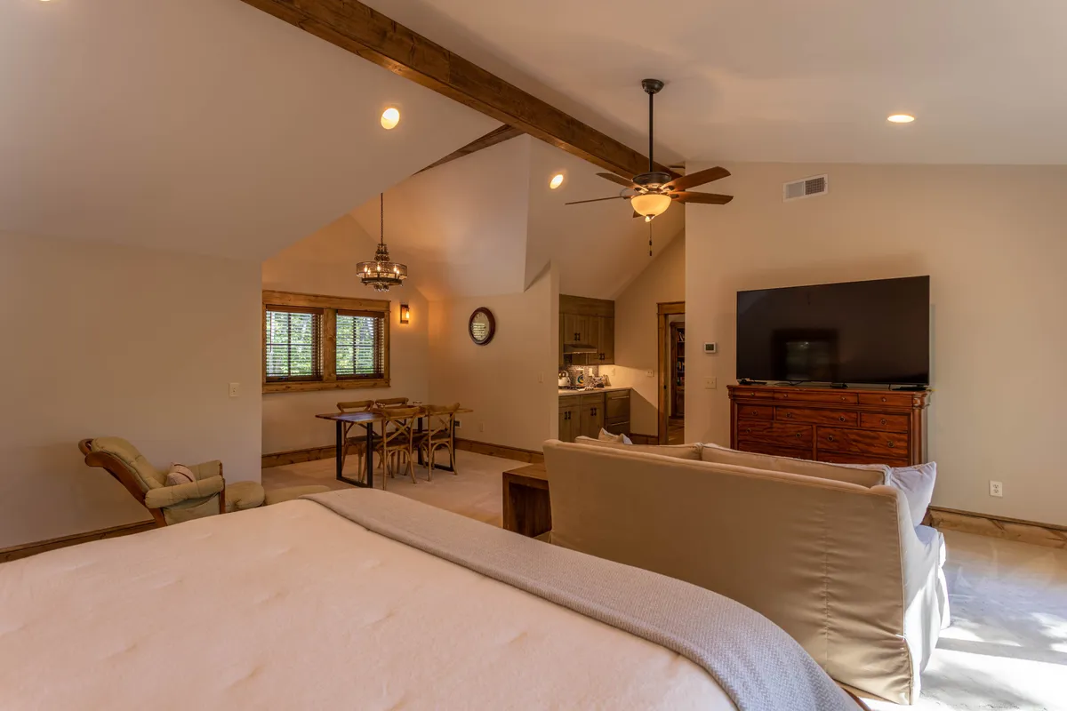 This image showcases a spacious and comfortable suite with a bed, sofa, dining table, and kitchen area, all under exposed ceiling beams. The space features warm, inviting decor and plenty of natural light, offering a sense of privacy and functionality. The inclusion of a kitchen area and seating highlights the versatility ideal for an in-law suite or master suite.