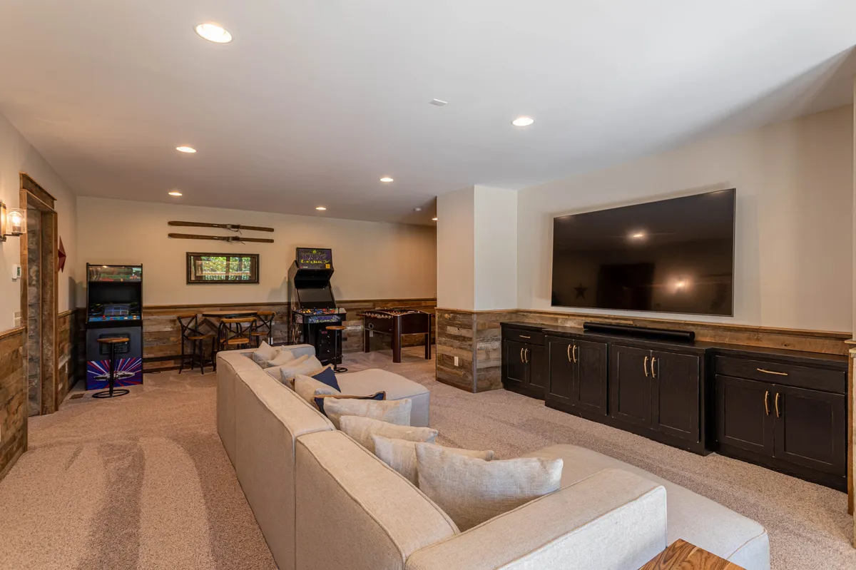 This image captures a spacious, lively game room featuring a comfortable sectional sofa, a large television perfect for movie nights, and a pair of arcade machines set against a wooden bar table. The ambiance is inviting and fun, providing plenty of space to relax or compete in friendly games.