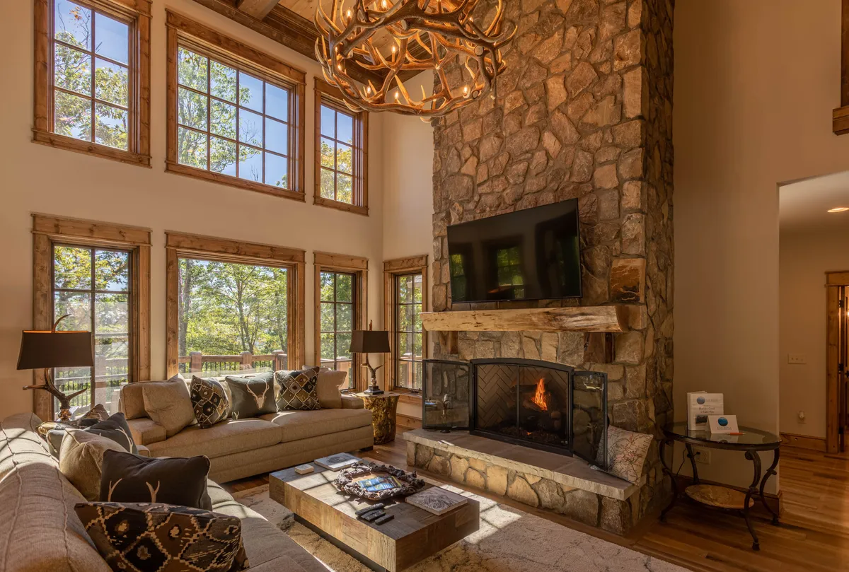 A spacious two-story Great Room with a dramatic stone fireplace, antler chandelier, large windows letting in natural light, a comfortable sofa, and a coffee table. The mountain views are visible through the windows, and the room feels cozy and inviting for gatherings.