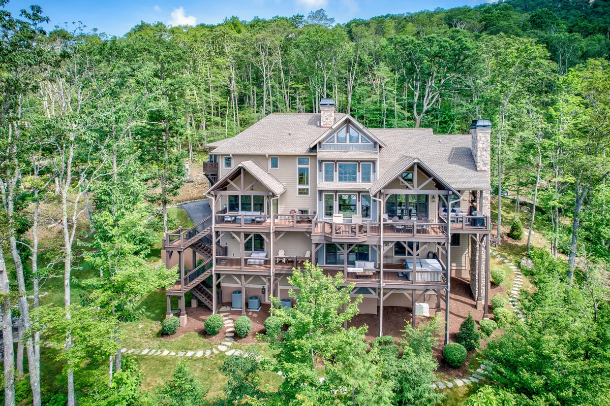 The image showcases a grand, multi-level custom mountain home surrounded by lush greenery. A stone pathway winds to the front entrance, while expansive balconies overlook the forested landscape and capture the warm glow of a sunset.
