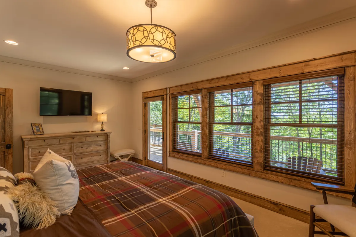 A master bedroom with a bed dressed in plaid bedding, large windows offering natural light and views, a wooden dresser, and a decorative lamp. The space is cozy and serene, emphasizing plush bedding and thoughtful decor.