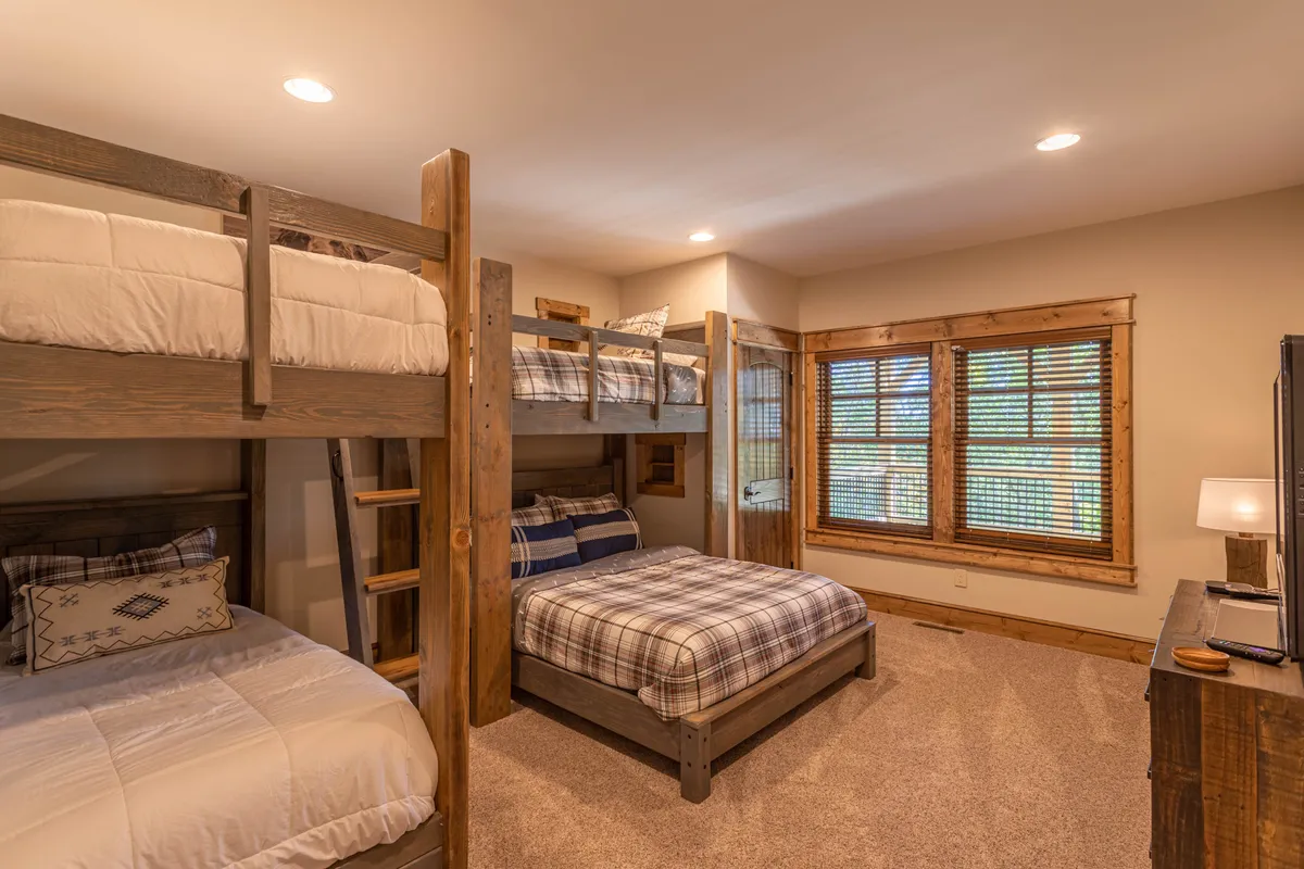 A bright and welcoming bunk room featuring custom-built bunk beds with plaid bedding, large windows, and wooden furniture. The scene suggests a playful yet restful environment ideal for children or groups, capturing the essence of fun and relaxation.